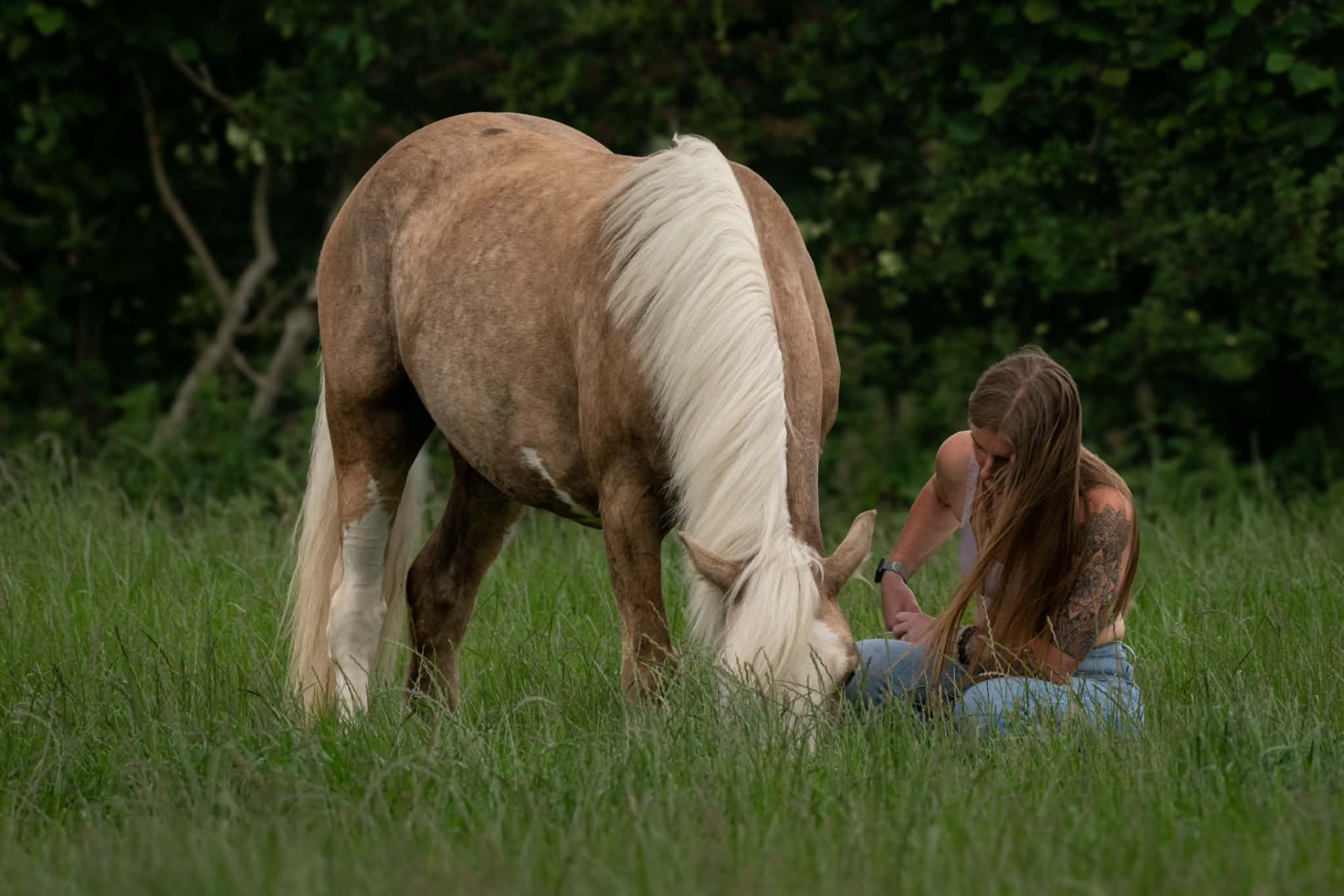 Woman sitting in long grass beside her grazing palomino pony