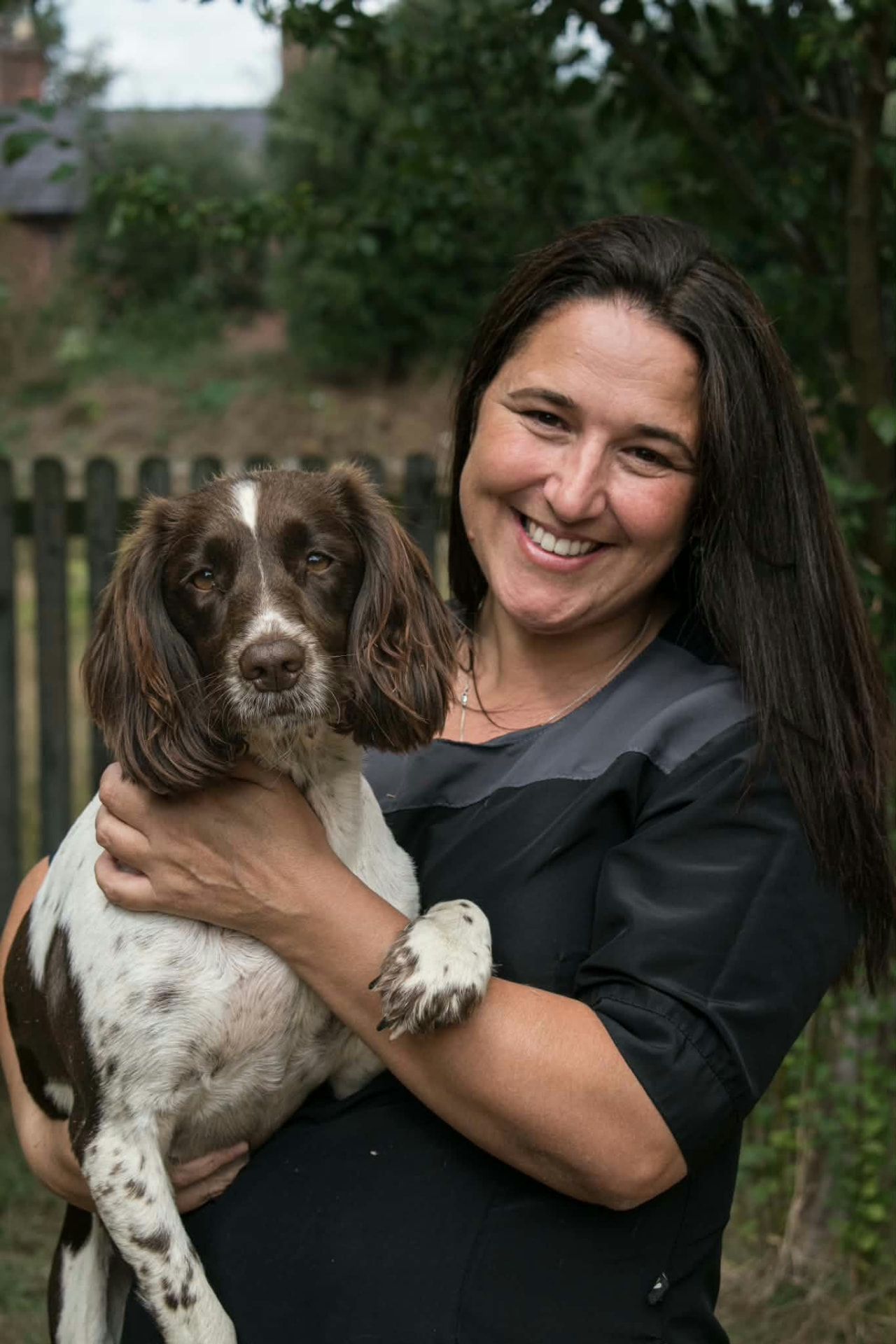 Smiling woman in black workwear holding her brown and white springer spaniel outdoors